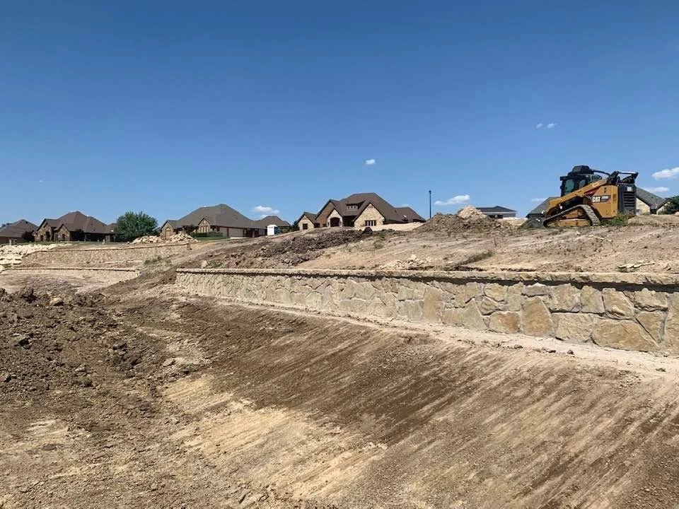 Excavated residential lot with stone retaining wall, graded earth, and CAT equipment on-site