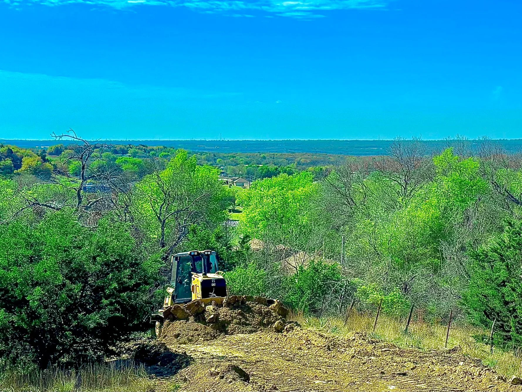 Excavator on cleared hillside with exposed soil, surrounded by green trees and brush