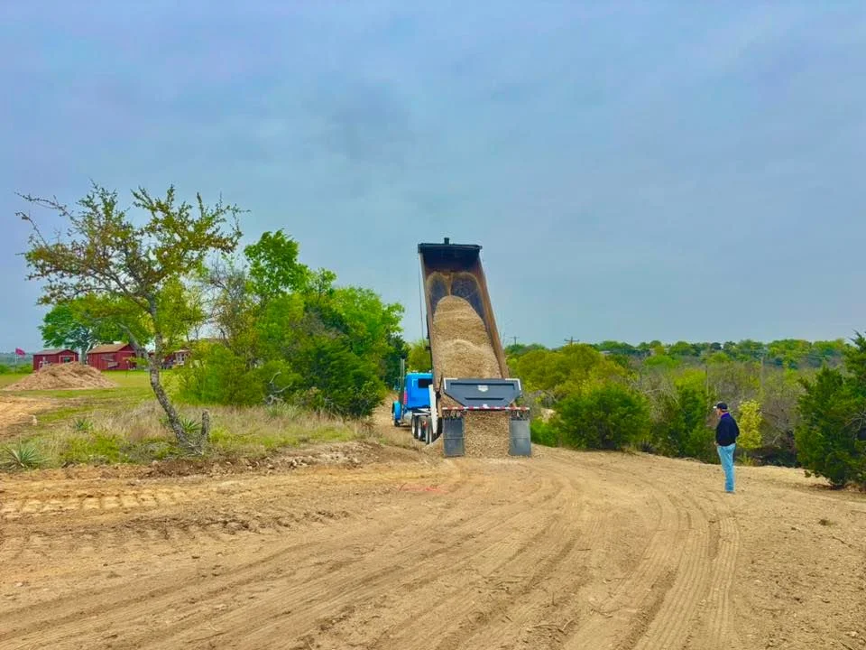 Dump truck unloading aggregate on graded driveway with compacted soil base