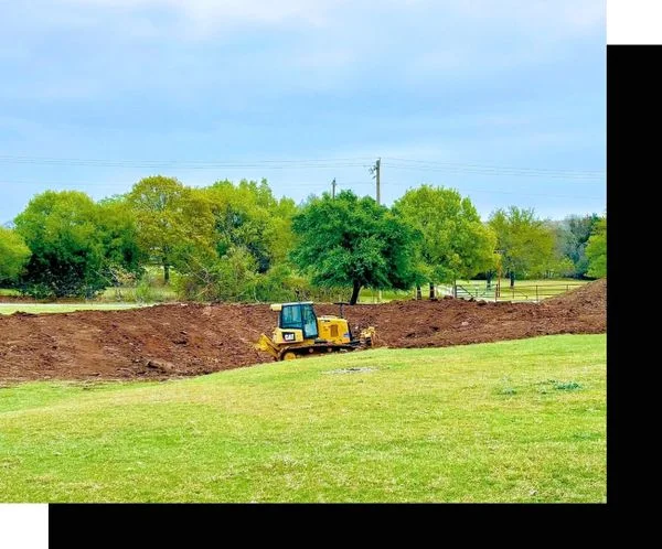 Yellow excavator grading and leveling dirt on residential property with mature trees in background